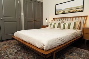 Une chambre avec un lit en bois, une table de chevet avec une lampe, une porte grise et un tableau représentant un paysage, accroché au mur.