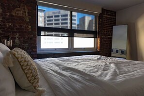 A hotel room with a bed, a window with a view of buildings, a brick wall, and a whiteboard on the wall. - Stay Roanoke | Luxury Brick Loft in Downtown Core (Roanoke)