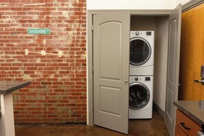 A laundry room with a brick wall, a door, and two stacked washing machines. - Stay Roanoke | Luxury Brick Loft in Downtown Core (Roanoke)
