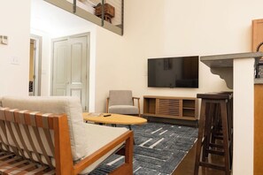 Uma sala de estar moderna com um sofá, uma mesa redonda pequena, uma TV fixada na parede e um armário de madeira.