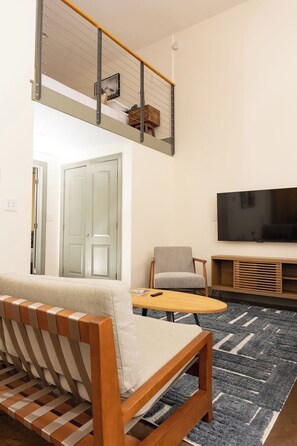 A modern living room with a sofa, a coffee table, a TV, and a staircase. - Stay Roanoke | Historic brick charm 1BR downtown (Roanoke)