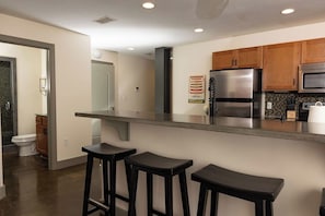 A modern kitchen with a stainless steel refrigerator, a microwave, and wooden cabinets. There are three black barstools in front of the counter. - Stay Roanoke | Downtown 2BR w/exposed brick charm (Roanoke)