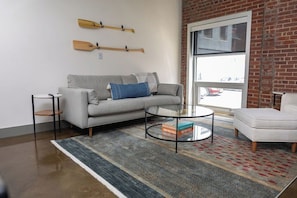 A modern living room with a grey sofa, a round glass coffee table, and a patterned rug. - Stay Roanoke | Downtown 2BR w/exposed brick charm (Roanoke)