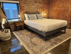 A bedroom with a brick wall, a bed with a grey frame, a patterned rug, a bedside table with a lamp, and a window with a view. - Stay Roanoke | Downtown 2BR w/exposed brick charm (Roanoke)