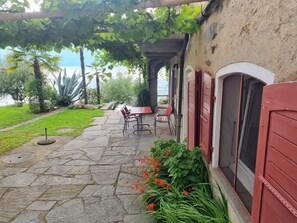Pátio de pedra com mesa e cadeiras, pérgola coberta por trepadeiras e vista para a água.