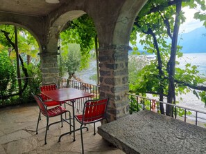 Eine Steinterrasse mit Tisch und Stühlen, mit Blick auf ein Gewässer und viel Grün.