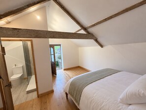 Une chambre avec un plafond en bois, un lit recouvert de linge de lit blanc et une vue sur un jardin par une porte ouverte.