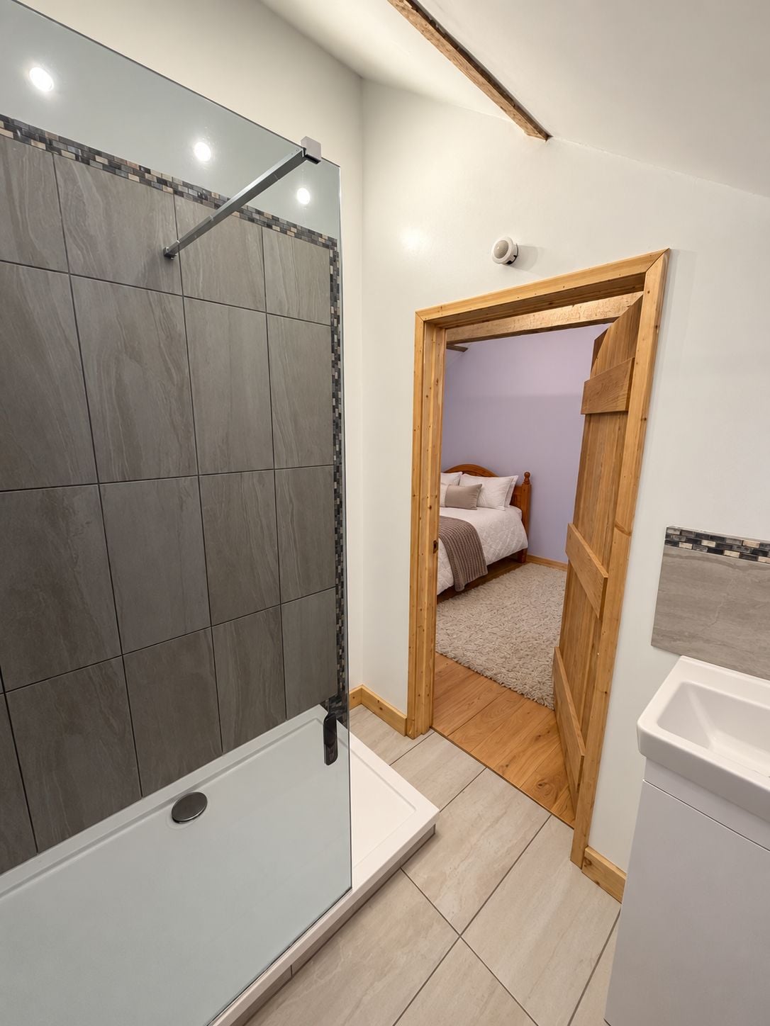 A bathroom with a shower, a toilet, and a sink. There is a doorway leading to a bedroom with a bed and a nightstand.