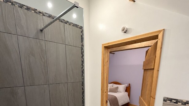 Une salle de bain avec une douche, des toilettes et un lavabo. Une porte donne sur une chambre à coucher qui contient un lit et une table de chevet.