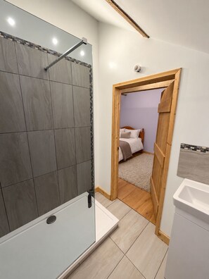 Bany amb dutxa, vàter i rentamans. Hi ha una porta que condueix a un dormitori amb un llit i una petita taula de nit.