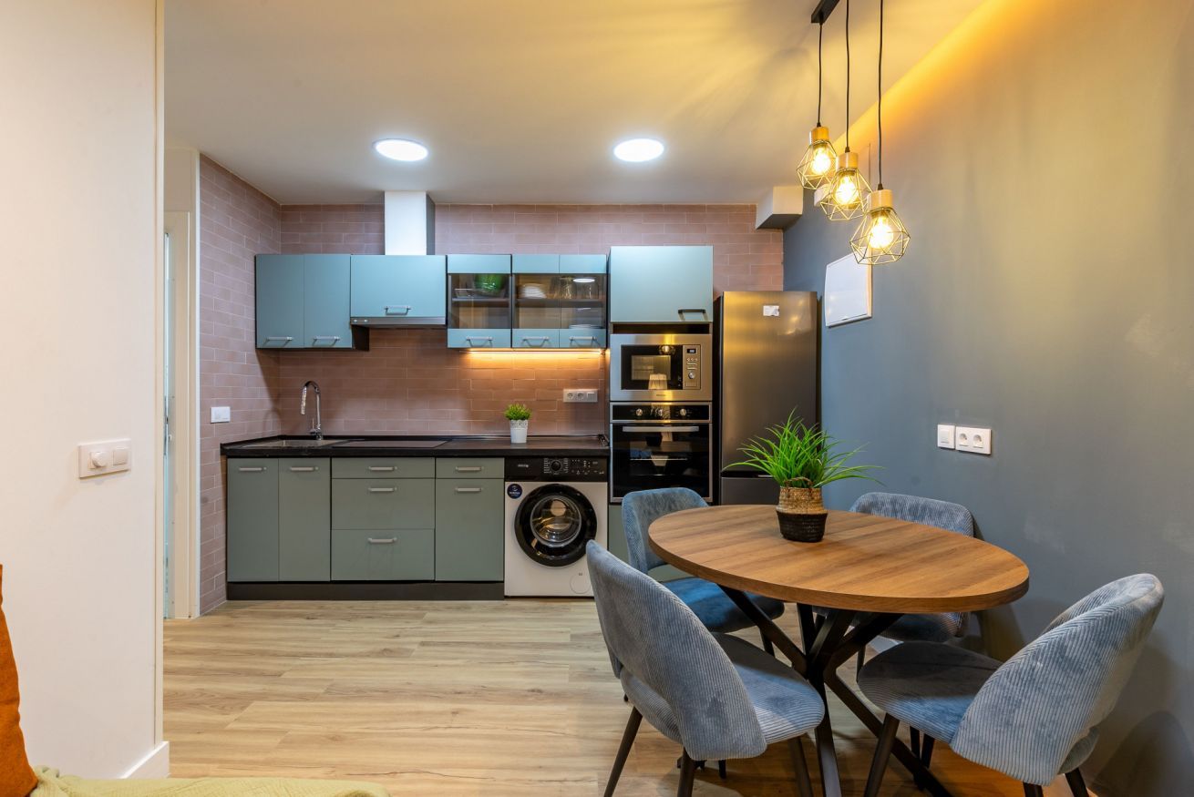 Una cocina moderna con armarios grises, horno empotrado, lavadora y comedor con una mesa redonda de madera y sillas tapizadas en gris.