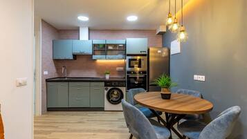 Una cocina moderna con armarios grises, horno empotrado, lavadora y comedor con una mesa redonda de madera y sillas tapizadas en gris.