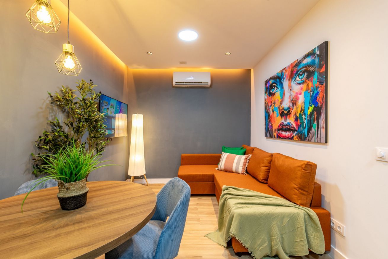 Una sala de estar moderna con una mesa de comedor de madera, sillas azules, un sofá naranja, una manta verde, una planta en maceta y un cuadro abstracto colorido.