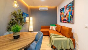 Ein modernes Wohnzimmer mit einem Holzesstisch, blauen Stühlen, einem orangefarbenen Sofa, einer grünen Tagesdecke, einer Topfpflanze und einem farbenfrohen abstrakten Gemälde.