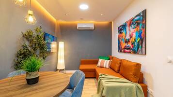 Una sala de estar moderna con una mesa de comedor de madera, sillas azules, un sofá naranja, una manta verde, una planta en maceta y un cuadro abstracto colorido.