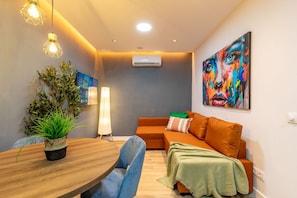 A modern living room with a wooden dining table, blue chairs, an orange sofa, a green throw blanket, a potted plant, and a colorful abstract painting. - Appartement 2 chambres moderne et calme - Marqués Vadillo (Madrid)