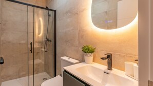 Ein modernes Badezimmer mit Duschkabine aus Glas, einem weißen Waschbecken und einem runden Spiegel sowie einer Topfpflanze.