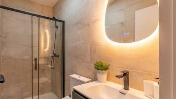 Un baño moderno con cabina de ducha de vidrio, un lavabo blanco con espejo redondo y una planta en maceta.