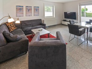 A modern living room with a grey sofa, a coffee table, and a TV. There are three abstract paintings on the wall, a window, and a radiator. - Holiday Apartment With 2 Bedrooms (Esens)