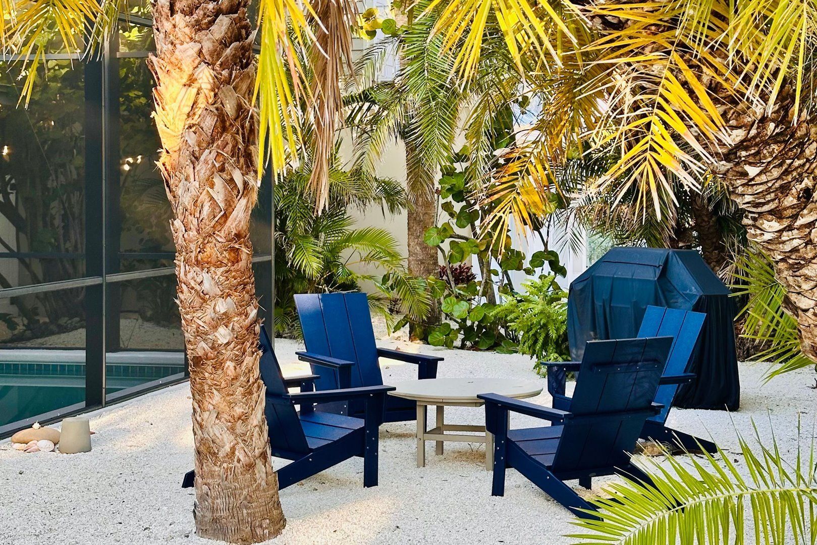 Two blue chairs and a small table in a garden area with palm trees and a pool.