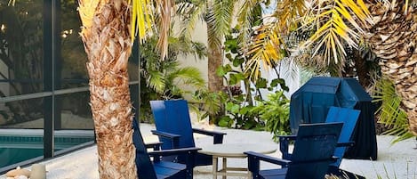 Two blue chairs and a small table in a garden area with palm trees and a pool.