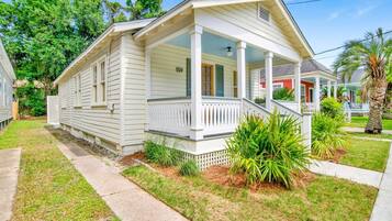 Uma casa térrea com varanda frontal, número de endereço visível e pequeno jardim da frente com grama e plantas.