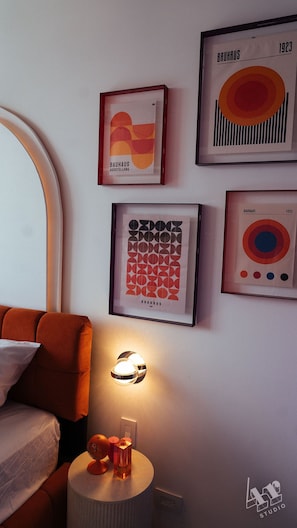 A room with a bed, a round side table with a lamp, and three framed artworks on the wall.
