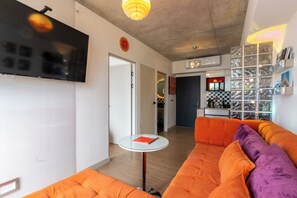 A modern living room with a large flat-screen TV, orange sofas, a small glass table, and a kitchen area in the background.