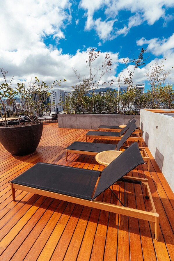 A rooftop terrace with wooden flooring, lounge chairs, a large planter, and a cityscape view.