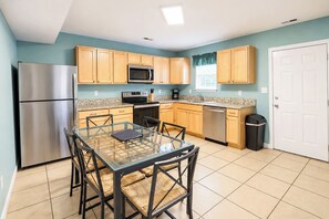 A modern kitchen with a stainless steel refrigerator, microwave, and dishwasher, wooden cabinets, a glass dining table with chairs, and a tiled floor. - Parrot Bay Townhouse 30 | Pet Friendly (Ocean City)