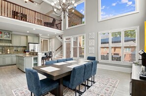 A spacious living area with a high ceiling, a dining table with blue upholstered chairs, a kitchen with light-colored cabinets, and a staircase leading to an upper level. - Hot Tub · Estate on the RTJ Fairway · Sleeps 16+ (Bessemer)