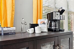 A coffee station with a coffee maker, cups, a spoon holder, and a sign for 'Wake Up Stays' in a hotel room. - Hot Tub · Estate on the RTJ Fairway · Sleeps 16+ (Bessemer)