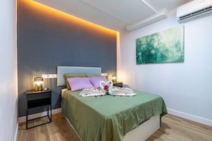 A neatly made bed with a green bedspread, two pillows, and a decorative swan made of towels. There is a nightstand with a lamp and a wall-mounted air conditioner. - Spacious and modern 2-bedroom apartment – Comillas (Madrid)
