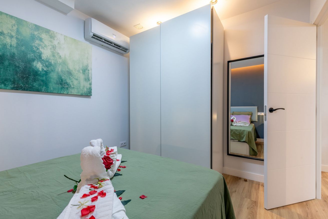 Habitación de hotel con cubrecama verde, toalla blanca con pétalos de rosa rojas, un espejo grande y un cuadro abstracto verde en la pared.