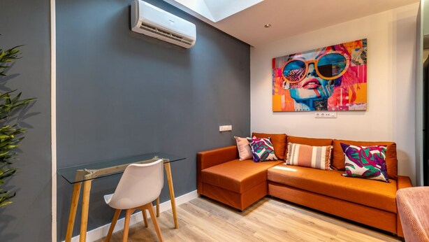 A modern living room with a glass table, a white chair, an orange sofa, and a colorful abstract painting. - Spacious and modern 2-bedroom apartment – ​​Comillas (Madrid)