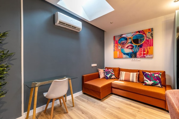 A modern living room with a glass table, a white chair, an orange sofa, and a colorful abstract painting. - Spacious and modern 2-bedroom apartment – Comillas (Madrid)