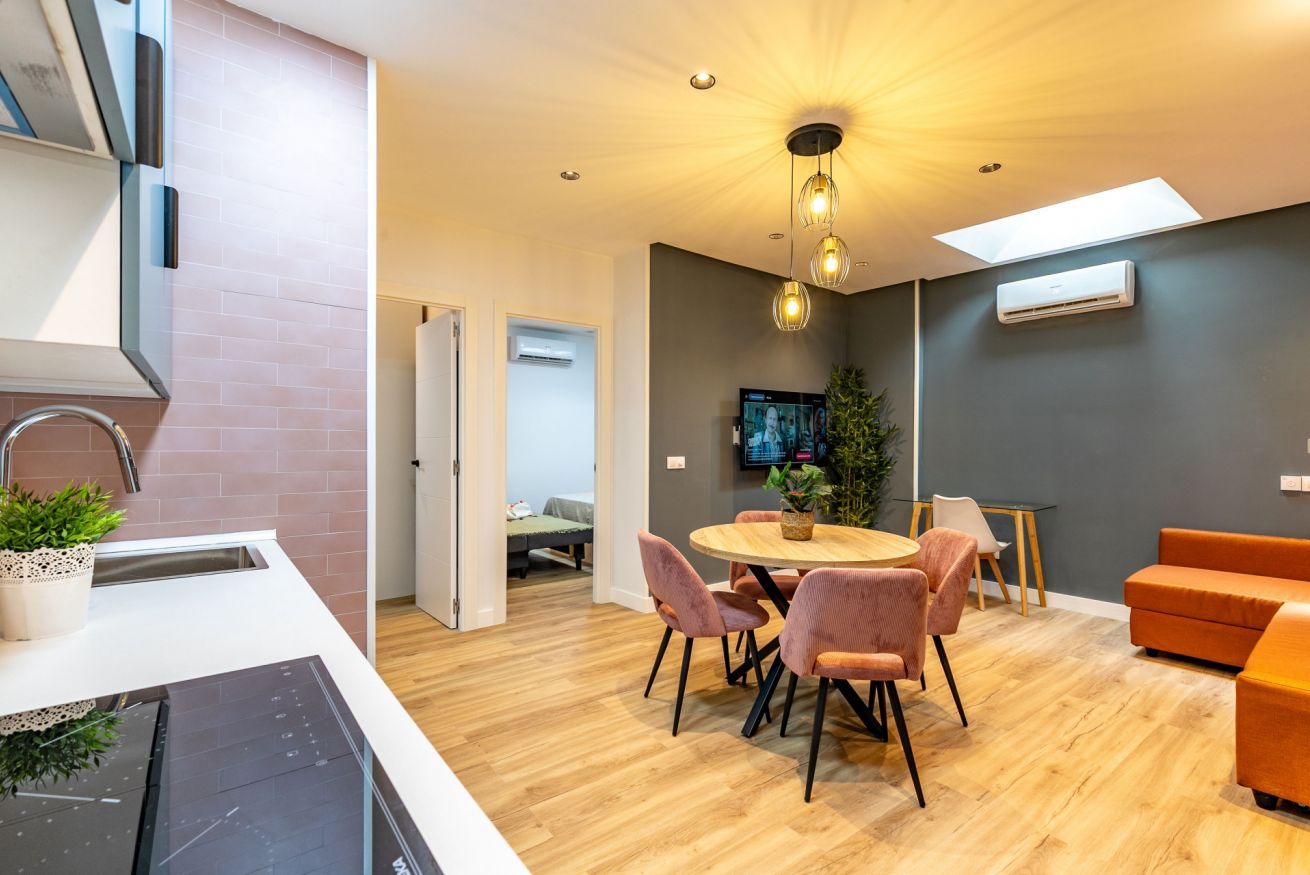 Una cocina moderna con estufa integrada, un comedor con mesa redonda y sillas, y una sala de estar con sofá y televisor.