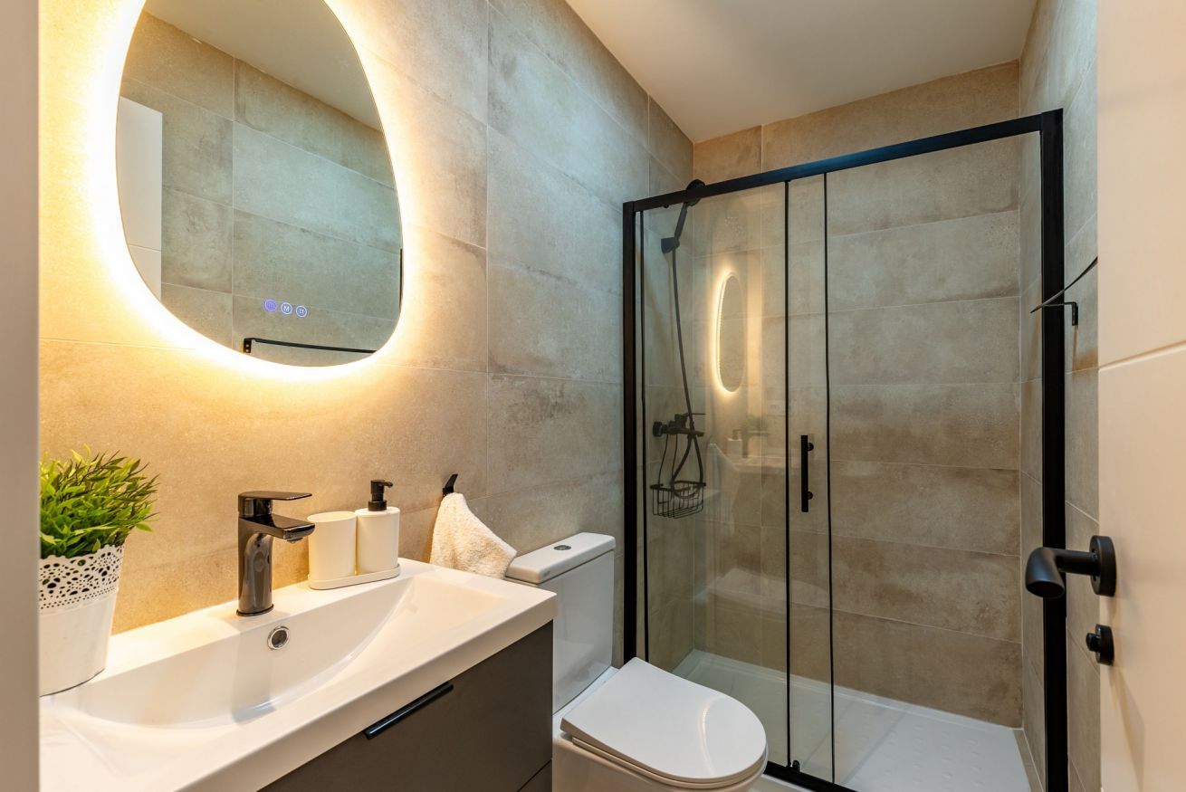 Un baño moderno con ducha de vidrio, un lavamanos blanco con espejo y una planta en maceta.