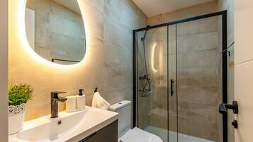 Un baño moderno con ducha de vidrio, un lavamanos blanco con espejo y una planta en maceta.