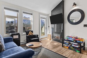 A modern living room with a fireplace, a sofa, a coffee table, and a collection of board games. - Mountain Views | Outdoor Heated Pool | Hot Tub (Canmore)