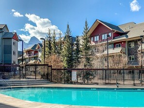 A swimming pool with a metal fence, surrounded by buildings with a mountainous backdrop. - Mountain Views | Outdoor Heated Pool | Hot Tub (Canmore)