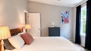 Camera da letto con un letto, lampade sui comodini, una ventola a soffitto, un comò, una finestra con tende e un quadro appeso al muro.