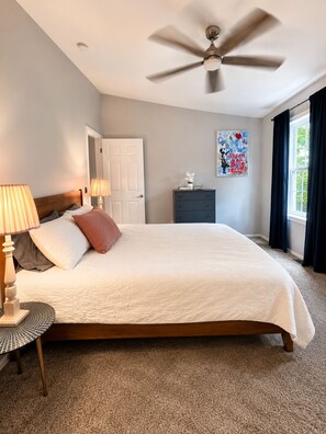 Un dormitorio con cama, lámparas de noche, ventilador de techo, cómoda, ventana con cortinas y una obra de arte en la pared.