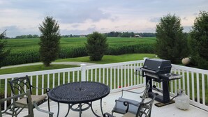 Una terraza con parrilla y asientos, con vista a un campo de maíz y un granero rojo.