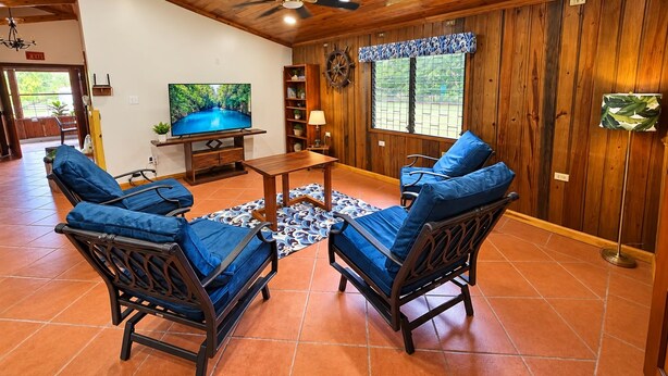 Une pièce aux murs et au plafond en bois, avec un téléviseur, deux fauteuils bleus rembourrés, une petite table et un tapis à motifs.