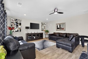 A modern living room with a sofa set, a flat-screen TV, decorative mirrors, and a ceiling fan. - Beautiful 5-bedroom house with all the amenities and convenient location. (Ellenwood)