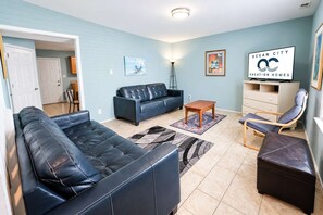 A living room with a blue couch, a wooden coffee table, a blue armchair, and a massage chair. There is a painting on the wall and a sign for Ocean City Vacation Homes. - Parrot Bay Townhouse 27 | Pet Friendly (Ocean City)