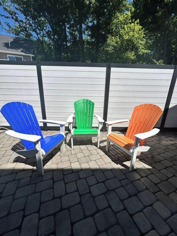 Tres sillas Adirondack en azul, verde y naranja, dispuestas en un patio empedrado con una valla blanca al fondo.