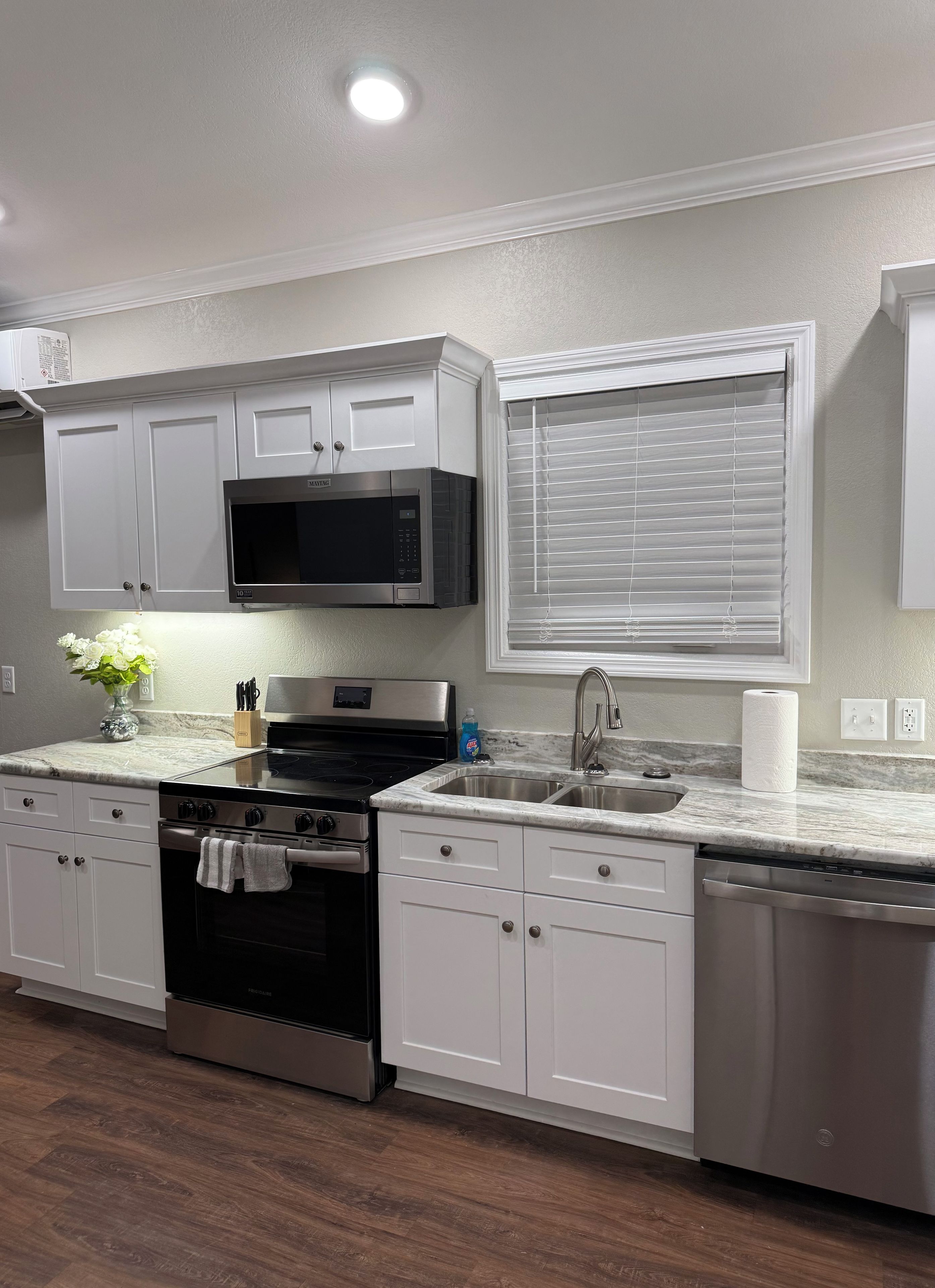 A modern kitchen with white cabinetry, a stainless steel microwave, a built-in oven, a dishwasher, and a window with blinds.