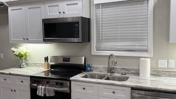 A modern kitchen with white cabinetry, a stainless steel microwave, a built-in oven, a dishwasher, and a window with blinds.
